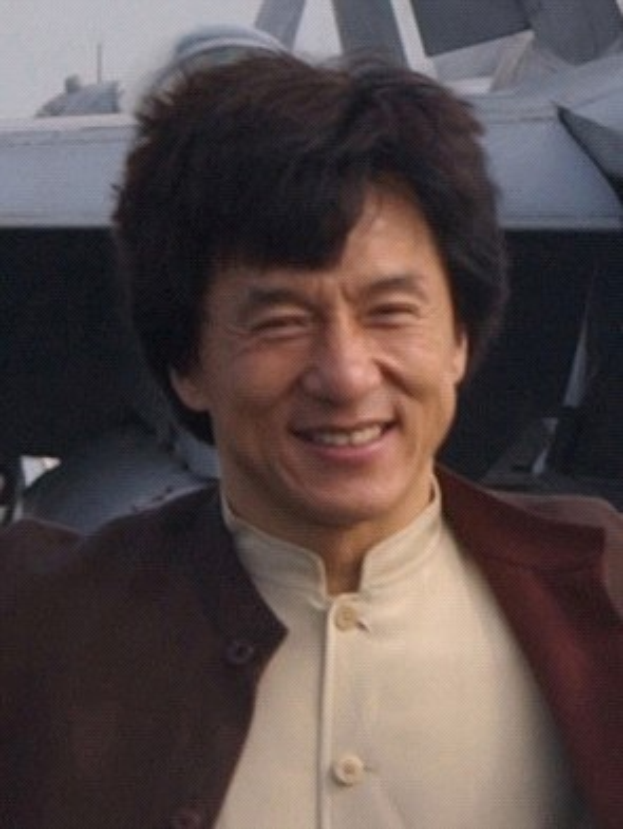 Jackie Chan steht lachend vor einem Flugzeug gegen einen klaren blauen Himmel, trägt ein weißes Hemd und eine schwarze Jacke mit ordentlich gekämmtem Haar.