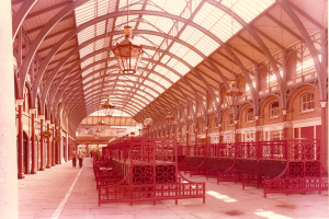 Rote Bänke in einem Bahnhof mit Glasdach, Menschen gehen, Geländer, Säulen, Deckenleuchten und ein Gebäude mit Fenstern im Hintergrund; Text erscheint unten.