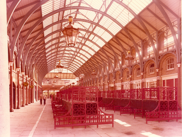 Rote Bänke in einem Bahnhof mit Glasdach, Menschen gehen, Geländer, Säulen, Deckenleuchten und ein Gebäude mit Fenstern im Hintergrund; Text erscheint unten.