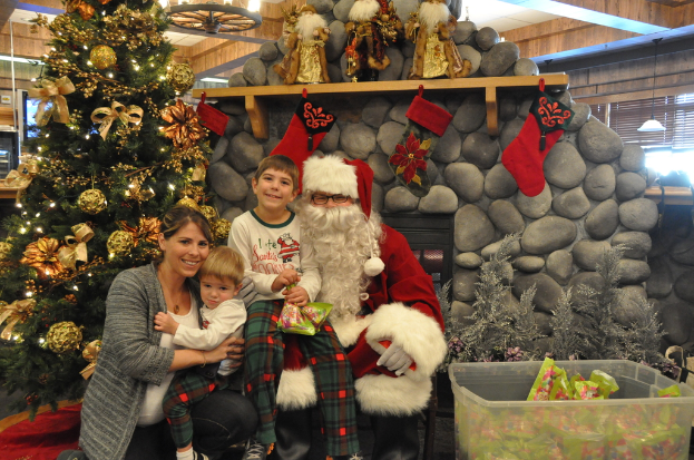 Eine Frau, Kinder und ein Mann in Santa-Claus-Kostüm mit Brille und Handschuhen stehen neben einem geschmückten Weihnachtsbaum, einer Wand mit Felsen, Spielzeugen, Dekorationen, einem Korb mit Gegenständen und Lichtern im Hintergrund. Ein Junge hält ein Päckchen.