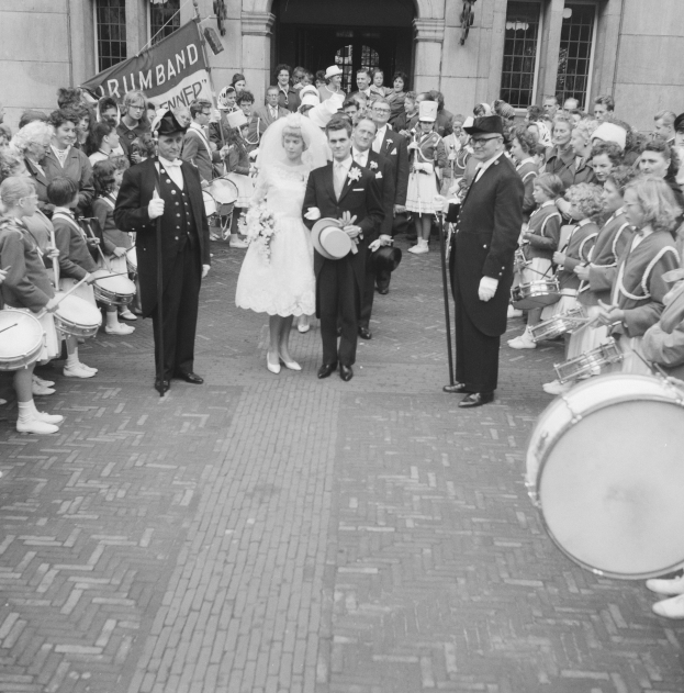 Eine Braut in einem weißen Kleid und Schleier und ein Bräutigam mit einem Blumenstrauß gehen eine Straße entlang vor einer Menge, einige mit Musikinstrumenten, mit einem Gebäude und einem Banner im Hintergrund, dargestellt in Schwarz-Weiß.