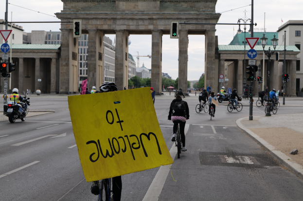 Eine Gruppe von Radfahrern mit Helmen fährt an der Brandenburgertor in Berlin vorbei, einer hält ein gelbes Schild, mit Laternenpfählen, Verkehrsampeln, Gebäuden, Bäumen und einem klaren blauen Himmel im Hintergrund.