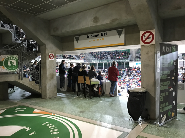 Eine Gruppe von Menschen steht vor einem sitzenden Publikum in einem Stadion mit Schildern, einem Müllbehälter, einer Texttafel, einer Treppe mit Geländern und Deckenleuchten; die Schilder tragen die Aufschrift "Boston Celtics vs New York Jets."