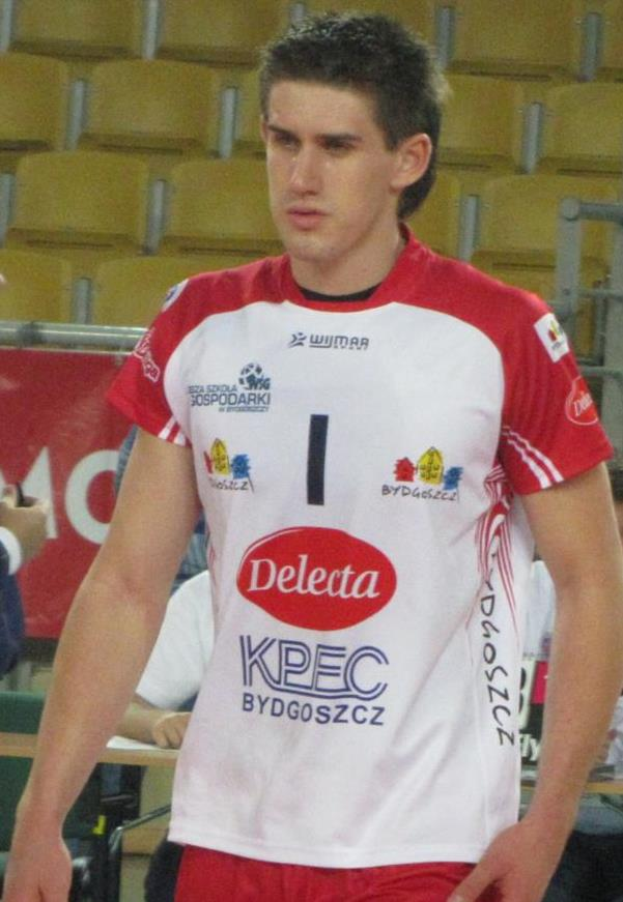 Ein Mann in einem roten und weißen Handballtrikot steht auf einem Feld, umgeben von Zuschauern, mit einem Banner im Hintergrund, das 'Krasnoyarsk vs Bydgoszcz' zeigt, und sieht entschlossen aus.