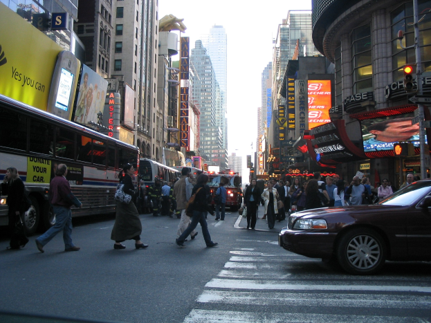 Eine belebte Straße mit Fahrzeugen, über die Straße gehenden Menschen, Gebäuden, Schildern, Verkehrsampeln und einem klaren blauen Himmel.