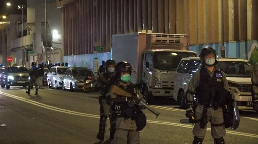 Eine Gruppe von Polizisten in Helmen, Handschuhen und Gesichtsmasken steht auf einer Straße bei Nacht, einige halten Waffen, mit Fahrzeugen, Laternenmasten, Schildern und Gebäuden im Hintergrund.