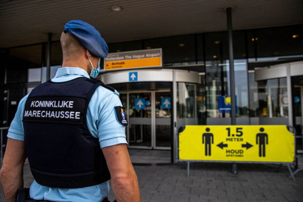 Polizist in blauer Uniform und Maske vor einem Glasgebäude mit mehreren Informationsschildern in der Nähe.