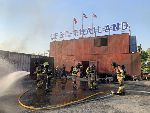 Feuerwehrleute in Helmen, mit Schläuchen, sprühen Wasser auf ein Gebäude mit Text, unter einem klaren blauen Himmel mit Fahnen und Bäumen im Hintergrund.