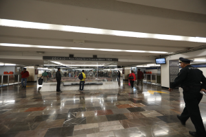 Eine Gruppe von Menschen durchquert eine belebte U-Bahn-Station, mit einem Polizeibeamten im Vordergrund, umgeben von Geländern, Bildschirmen und Texttafeln an der Decke.