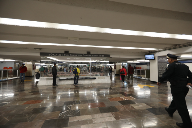 Eine Gruppe von Menschen durchquert eine belebte U-Bahn-Station, mit einem Polizeibeamten im Vordergrund, umgeben von Geländern, Bildschirmen und Texttafeln an der Decke.