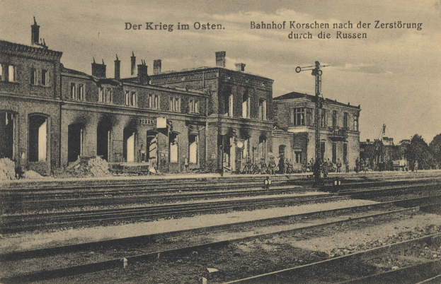Schwarzes und weißes Foto des Bahnhofs Korschen in Ostern, Deutschland, zeigt Gleise, Gebäude, Menschen, Bäume und Himmel mit Text oben und unten.