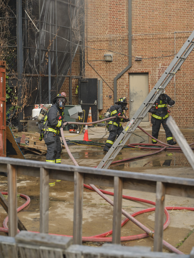 Feuerwehrleute in Helmen arbeiten daran, ein Gebäude Feuer zu löschen, während sie Schläuche halten, mit einem Metallzaun, Rohren, einem Container, einer Verkehrskegel und einem Baum im Hintergrund.