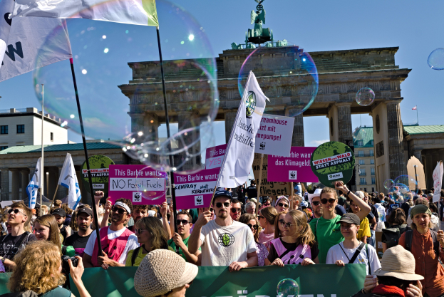 Eine Menge steht vor dem Reichstag in Berlin, viele tragen Mützen und Schutzbrillen, einige halten Fahnen und Schilder mit Text, mit Gebäuden, einem Tor, das von einer Statue gekrönt wird, und einem Himmel voller Seifenblasen im Hintergrund.