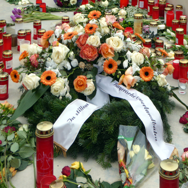 Ein Denkmal in Berlin, Deutschland, mit einem Blumenstrauß und Kerzen, die auf dem Boden vor einer Wand aufgestellt sind, umgeben von Blättern und anderen Gegenständen.