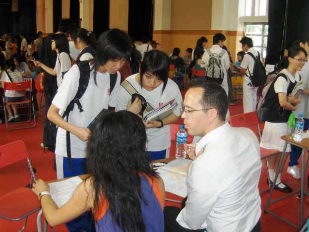 Ein Klassenzimmer mit mehreren Mädchen in weißen T-Shirts, die Bücher in der Hand halten, vorne, ein Mann und eine Frau im Vordergrund sitzend und diskutierend, und eine gelbe Wand mit einem Glasfenster im Hintergrund.