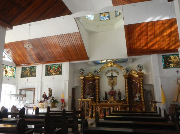 Innere einer Kirche mit Holzbankreihen, Statuen, Blumenbouquets, Fahnen, gerahmten Fotos an den Wänden und Buntglasfenstern, mit Deckenleuchten, die eine gemütliche Atmosphäre schaffen.