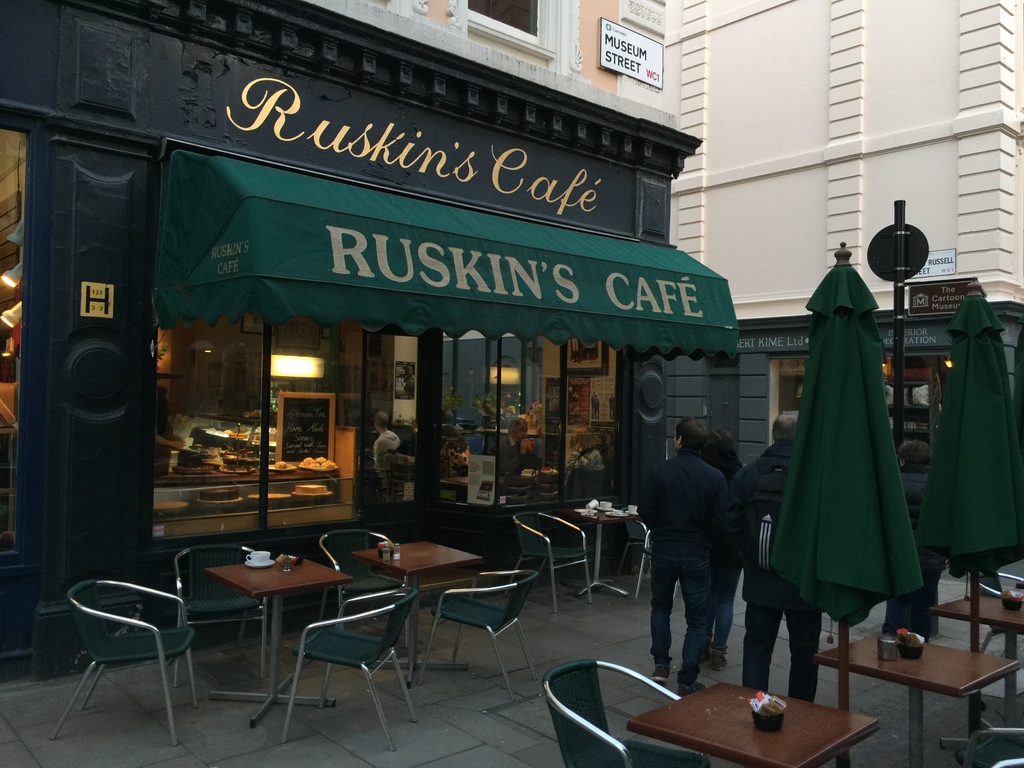 Eine Gruppe von Menschen steht vor einem Café mit einer Tafel, auf der 'Ruskins Café' steht.