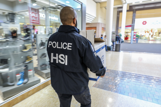 Polizeibeamter mit einer Kiste in einem Einkaufszentrum mit anderen Menschen im Hintergrund, der eine schwarze Jacke mit Text trägt, in der Nähe von Schuhregalen, Säulen, einem Mülleimer und Deckenlampen.
