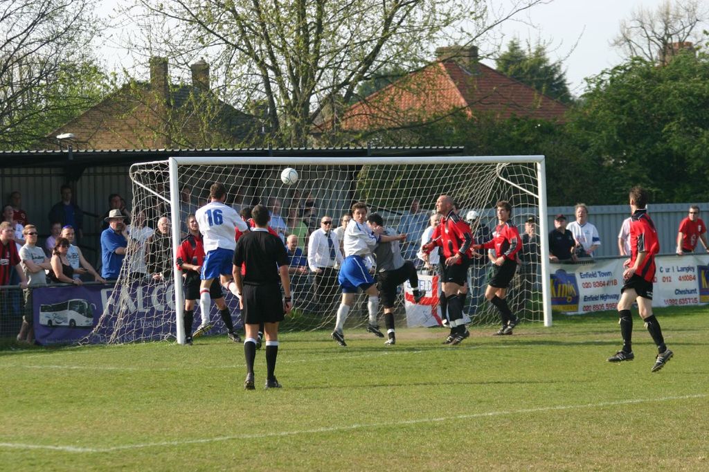 Fußballspieler spielen auf einem Feld mit einem Tor und Zuschauern hinter dem Netz, mit Bäumen und Häusern im Hintergrund.