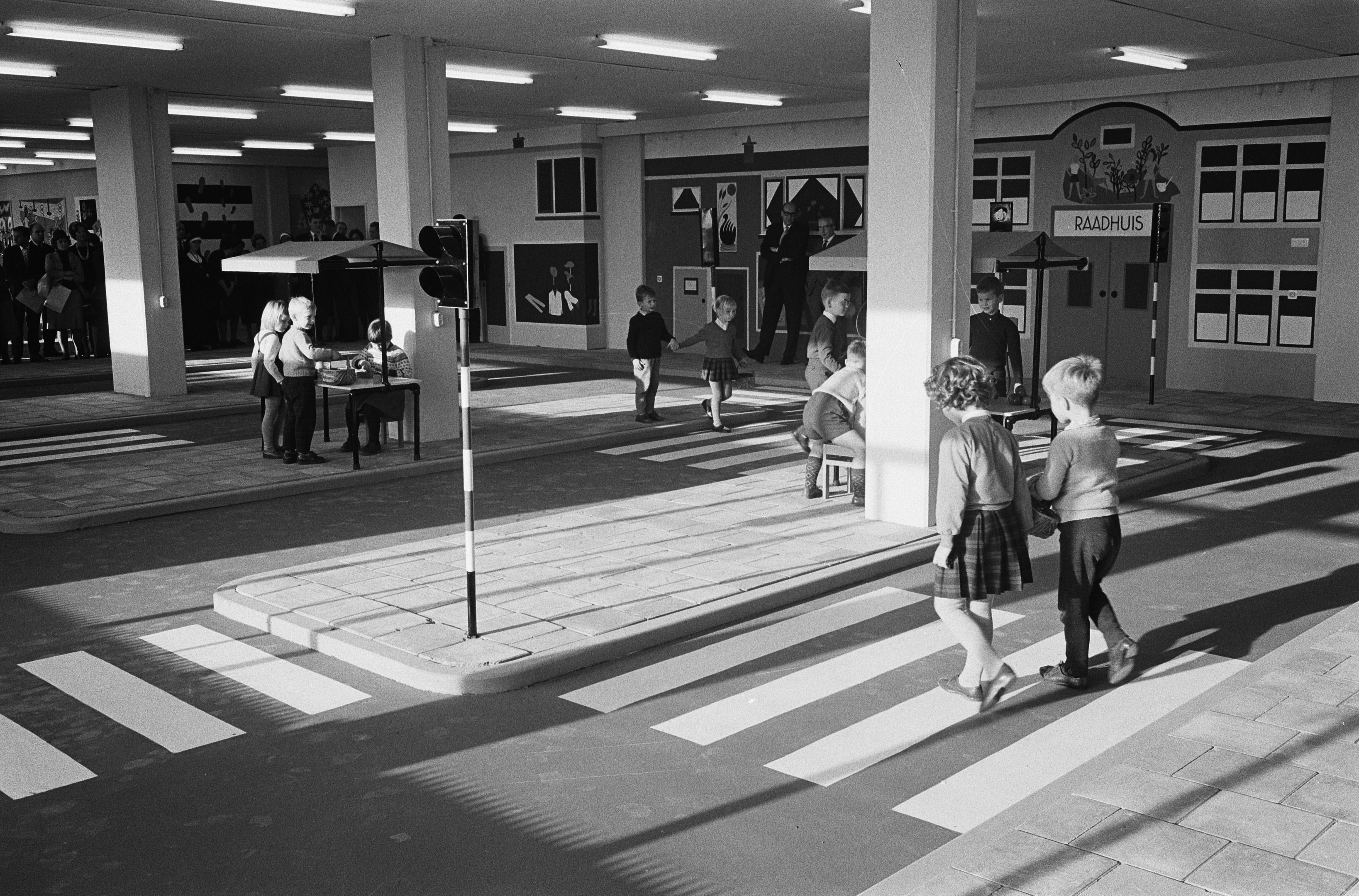 Schwarz-weiß-Foto von Kindern, die eine Straße an einer Ampel überqueren, mit Fußgängern in der Nähe, einem Verkehrszeichen im Vordergrund und strukturellen Pfeilern und Lampen im Hintergrund.