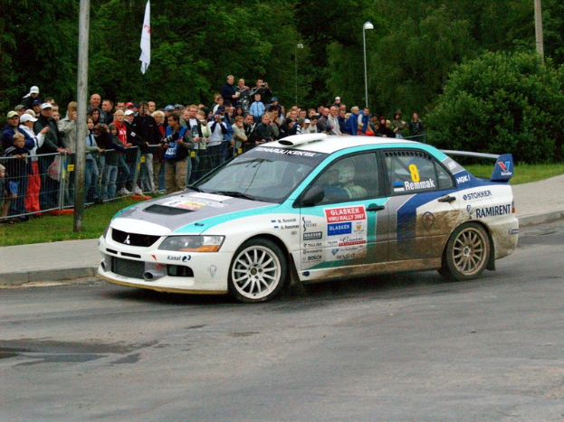 Ein Rallyeauto mit Text und Logos fährt an einer Menge vorbei, die Kameras hält, eine Straße entlang, die von Geländern, Polen mit Fahnen und Bäumen gesäumt ist.