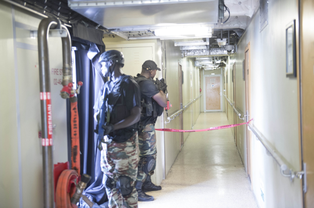 Zwei Polizisten in Uniform stehen nebeneinander in einem Flur, tragen Helme und halten Waffen, mit einer Tür und einem Rahmen auf der rechten Seite und Deckenleuchten und Rohren.