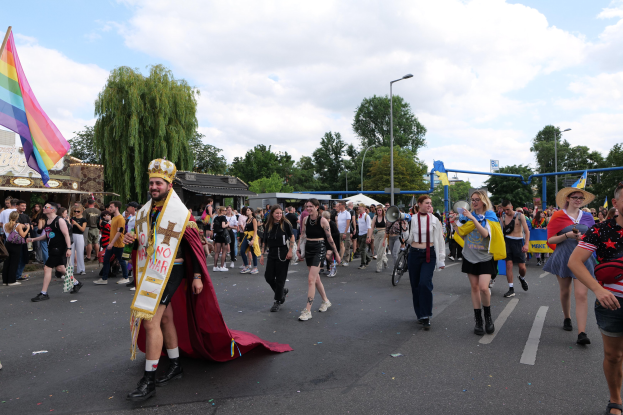 Eine Gruppe von Menschen bei der Gay Pride Parade 2018 mit einer Regenbogenflagge und Musikinstrumenten, einige tragen Mützen, vor dem Hintergrund von Laternenmasten, Bäumen, Schuppen und einem bewölkten Himmel.
