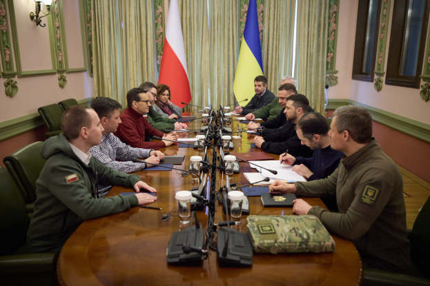 Gruppe von Männern in formeller Kleidung, die um einen Holztisch mit Mikrofonen und Dokumenten sitzen und in einem Raum mit Flaggen, Vorhängen und an der Wand montierten Leuchten diskutieren.