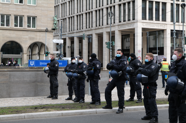 Eine Gruppe von Polizisten in schwarzen Uniformen und Masken steht vor einem Gebäude mit Glasfenstern und Säulen in Berlin, Deutschland, einige halten Helme, mit Laternen, Verkehrsampeln, Schildern mit Text, einer Statue und anderen Menschen im Hintergrund auf einer grünen Fläche.