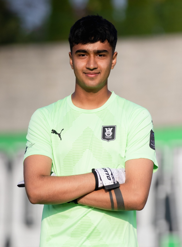 Ein junger Mann in einem grünen T-Shirt steht auf einem Fußballfeld und hat die Arme verschränkt, während er lächelt.