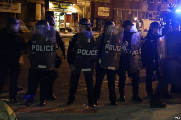 Polizisten in Helmen und Schilden stehen in einer Straße bei Nacht, mit Gebäuden, Fahrzeugen und Laternen im Hintergrund.
