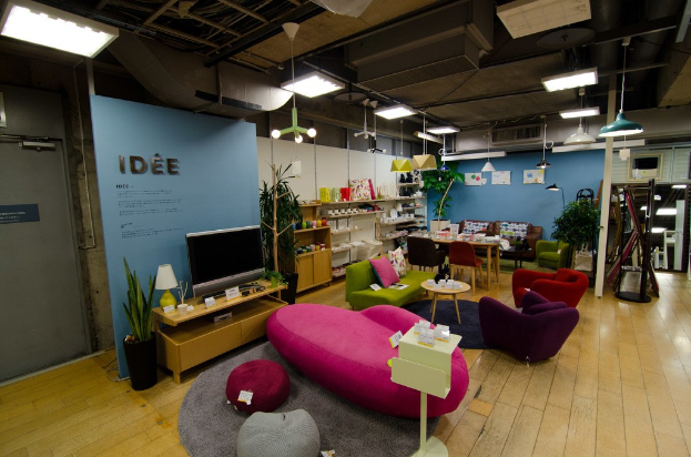 Haushaltsgeräte und Möbel sind in einem Geschäft wie in einem Wohnzimmer angeordnet, mit einer pinken Couch, Fernseher auf einem Tisch, Essbereich, Regalen mit Gegenständen und Deckenbeleuchtung.