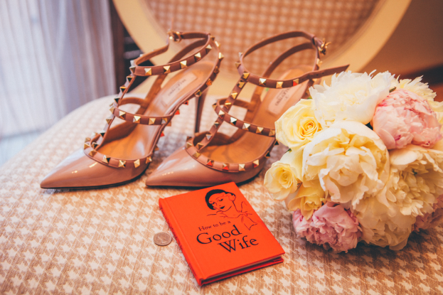Ein Tisch zeigt ein Buch mit dem Titel "Wie man eine gute Ehefrau ist", einen Blumenstrauß und ein Paar Valentino-Schuhe, vor einem Vorhang als Hintergrund.