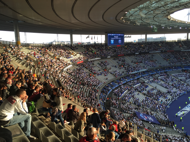 Eine große Menge Menschen sitzt in einem Stadion und schaut einem Fussballspiel zu, mit einer Bühne auf der rechten Seite, Fahnen, Masten und einem Bildschirm im Hintergrund, unter einem sichtbaren Himmel, im Allianz Stadion in München, Deutschland.