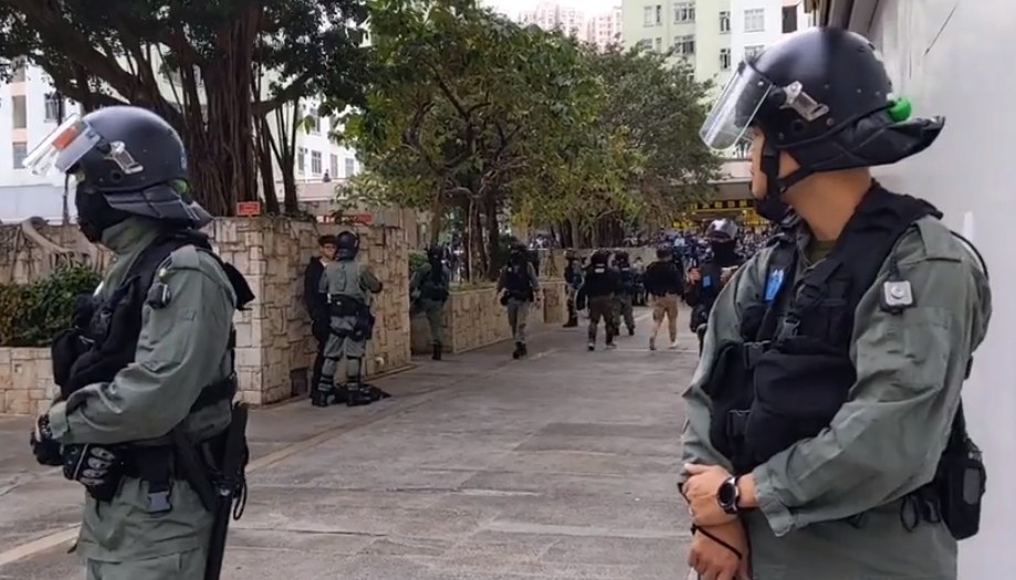 Polizisten in Helmen und kugelsicheren Westen stehen in einer Reihe auf einer Straße in Hong Kong, mit Bäumen, Gebäuden und Himmel im Hintergrund.