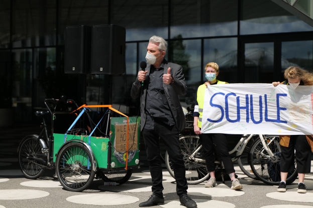 Ein Mann mit Maske spricht in ein Mikrofon auf einer Demonstration, hält ein Banner mit teilweise lesbarem Text, während andere in der Nähe stehen und ein weiteres Banner halten, mit Fahrrädern, einem Fahrzeug, Lautsprechern und einem glaswandigen Gebäude im Hintergrund.