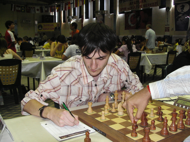A man's hand moves a chess piece on a board while writing with a pen, with people playing in the background.