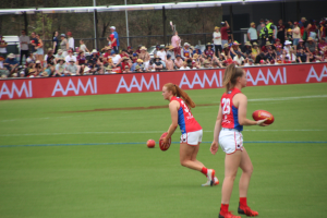 Zwei Frauen in Sportbekleidung beim Australian Football auf einem Feld, eine hält den Ball, mit Zuschauern und Werbetafeln im Hintergrund.