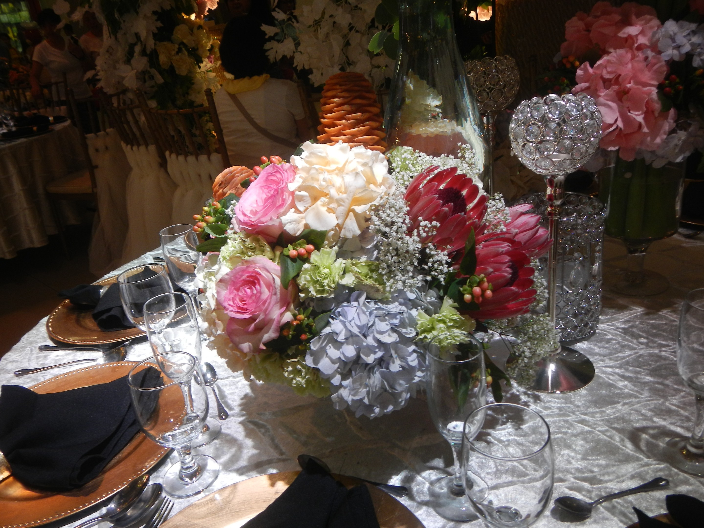 Ein formelles Dinner-Tisch-Arrangement mit Tellern, Besteck, Gläsern, Servietten und Blumenvasen, das eine florale Tischmitte und im Hintergrund Stühle und besetzte Tische zeigt.