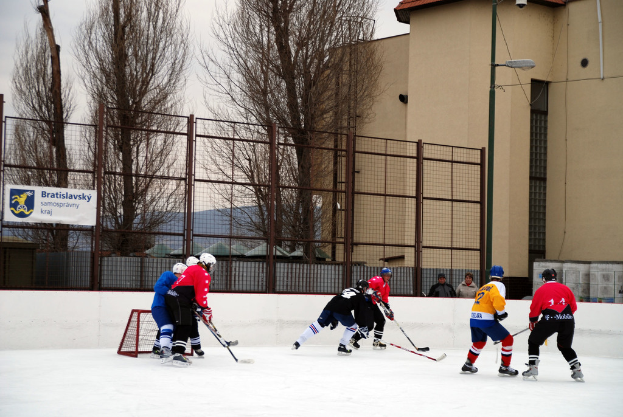 Personen beim Eishockey auf einem Eis mit Himmel, Bäumen, Gebäuden, Straßenlaterne, Namensschild und Zäunen im Hintergrund.