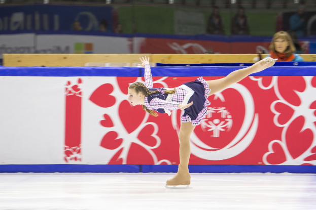 Eine Frau führt eine Eistanze auf einem Eisstadion aus, die Arme ausgestreckt, mit einem Kleid und Schlittschuhen bekleidet, während im Hintergrund Zuschauer und eine Tafel mit Text zu sehen sind.