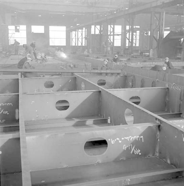 Ein Schwarz-Weiss-Foto einer Fabrik mit Arbeitern in Helmen, die an Stahlträgern arbeiten, mit Fenstern, Pfählen, Lampen und anderen Gegenständen im Hintergrund und Text unten.