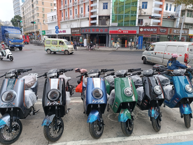 Eine Reihe von Elektro-Scootern, die entlang einer Straße geparkt sind, mit einer Person in einer Mütze davor, vor einem Hintergrund von Gebäuden, Verkehrsampeln, Bäumen und einem klaren Himmel.