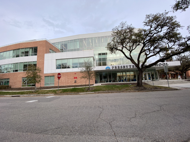 Großes zweistöckiges Backsteingebäude mit der Aufschrift "Presbyte Mittelschule" und vielen rechteckigen Fenstern, umgeben von Bäumen, Gras, Schildern, Strommasten und einer asphaltierten Straße unter einem bewölkten Himmel.