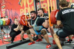 Eine Gruppe von Männern führt Kniebeugen mit Hanteln in einem mit Geräten und Bannern gefüllten Fitnessstudio durch, auf denen "Powerlifting-Weltmeisterschaften 2019" steht.