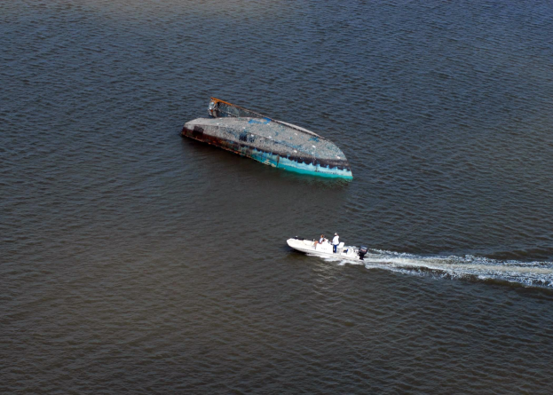 Ein kleines Boot fährt einen Fluss hinunter neben einem teilweise gesunkenen, havarierten großen Boot mit ein paar Menschen an Bord.