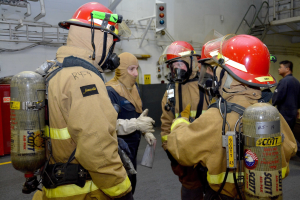 Feuerwehrleute in Helmen und Gasflaschen stehen in einem Raum mit Rohren, einer Ampel und einer Treppe mit Geländern im Hintergrund.