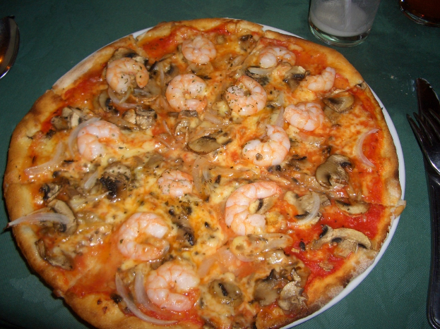 Eine Pizza mit Pilzen, Garnelen und anderen Belägen auf einem Teller, begleitet von einer Gabel, einem Messer, zwei Gläsern und einem Löffel links.