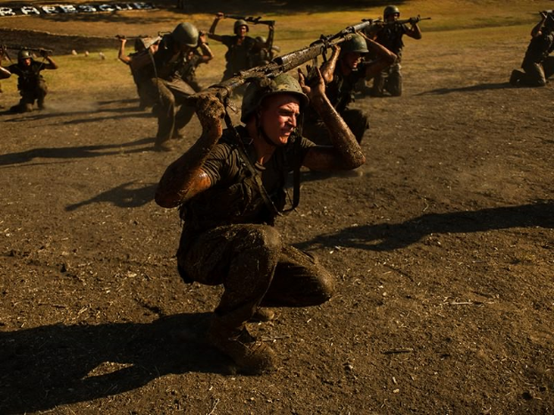Eine Gruppe von Männern in Militäruniformen, Helmen und mit Gewehren rennt über ein Feld mit Bäumen und Gras im Hintergrund und Text am unteren Bildrand.
