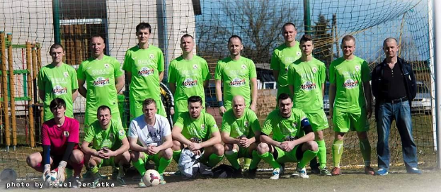 Gruppe von Männer in grünen T-Shirts auf einem Fussballfeld mit einem Ball und einem Torpfosten, umgeben von Bäumen, Geb&aumluden und Fahrzeugen.